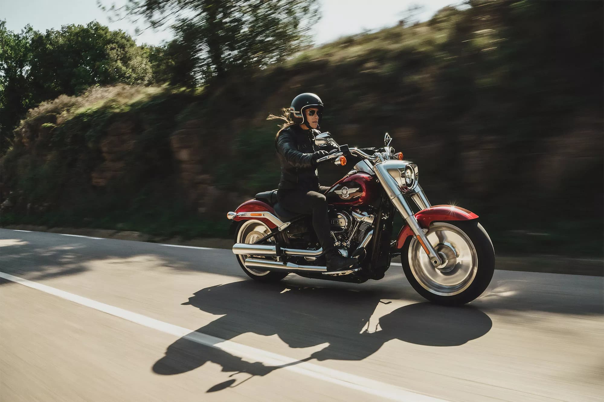 Rider on a red Harley-Davidson cruiser speeding past a sunlit hillside