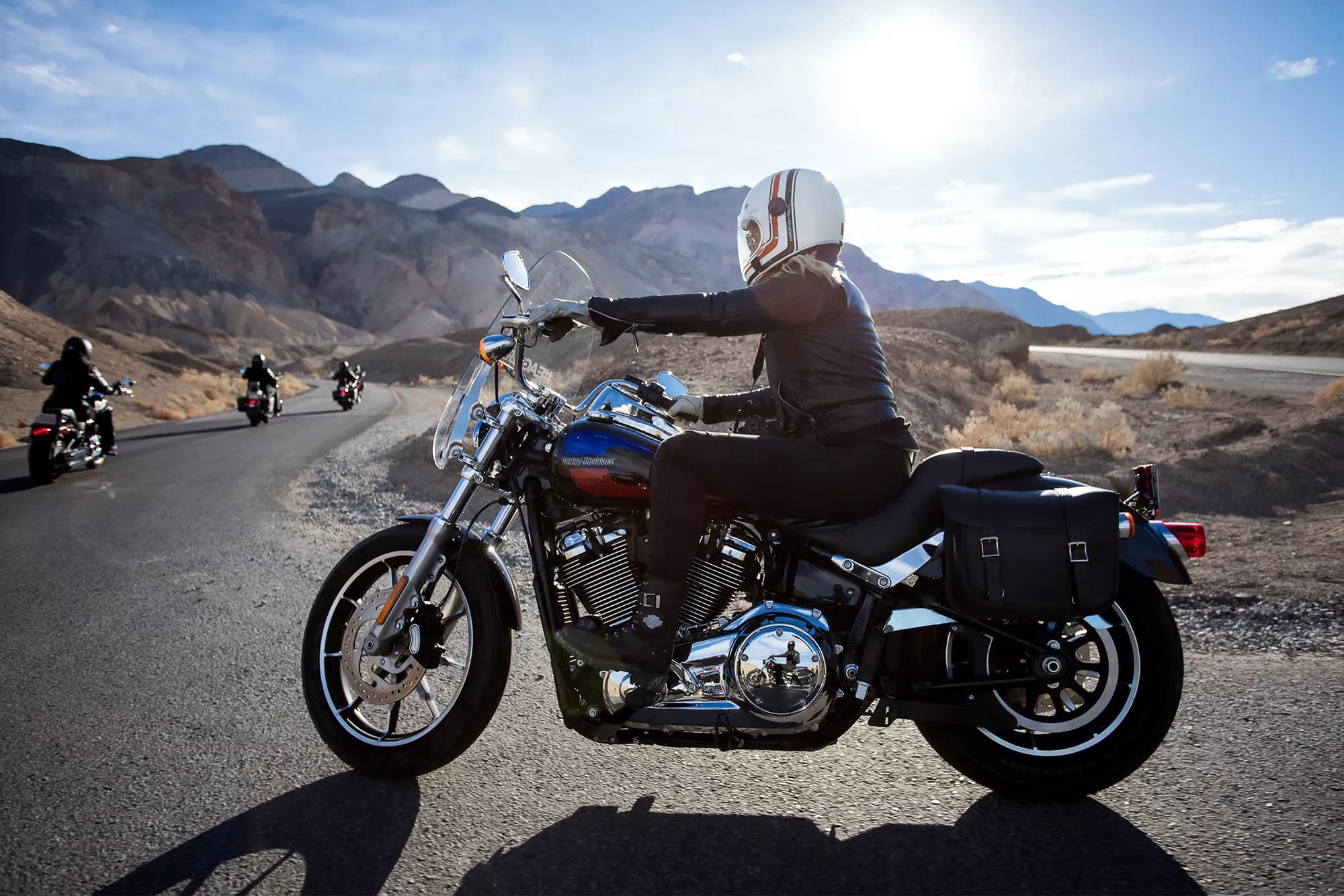 Riders on Harley-Davidson motorcycles on a mountain road under a bright sky